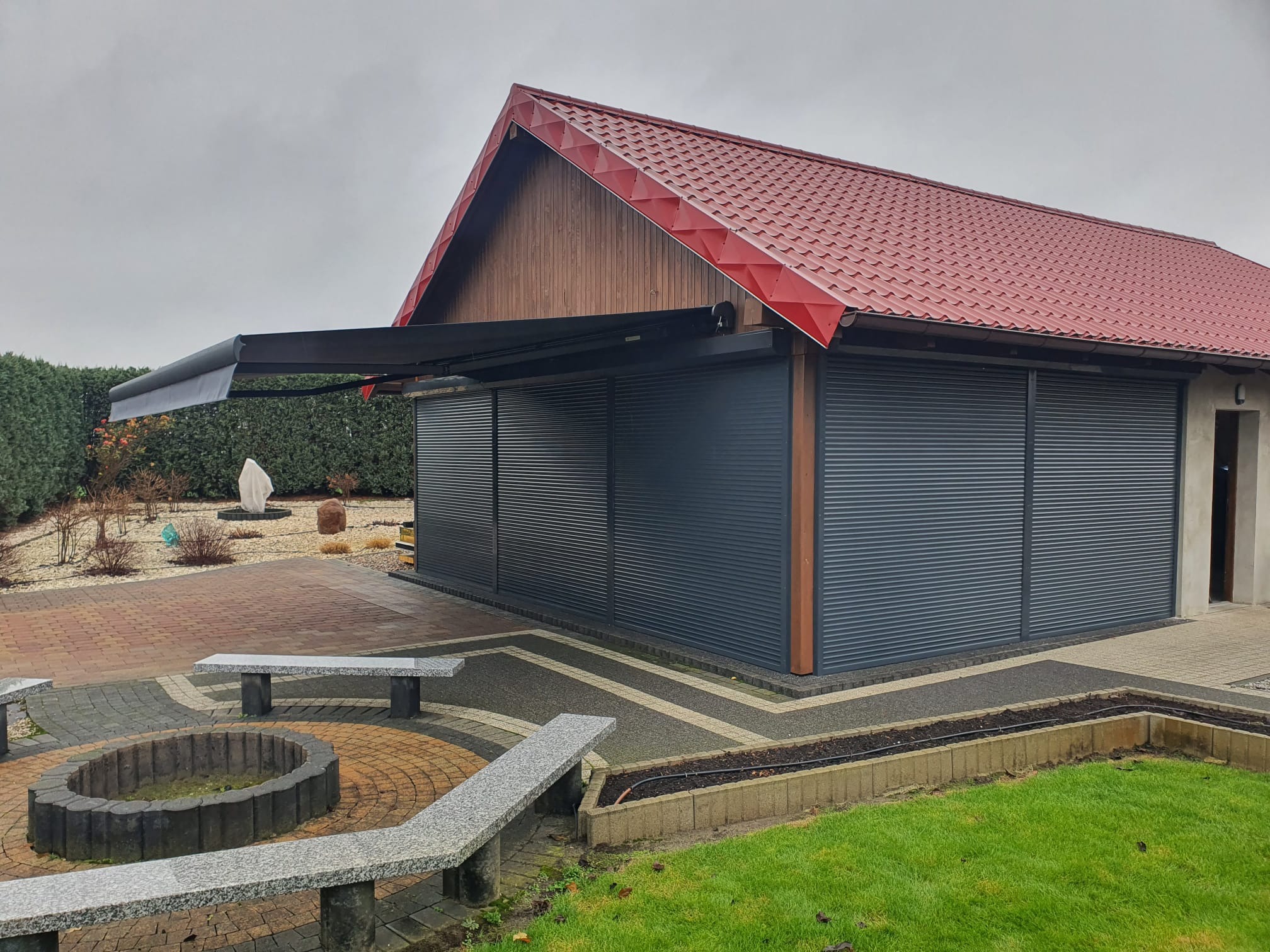 Zadaszone miejsce do wypoczynku z szarymi roletami zewnętrznymi, wysuniętą czarną markizą i czerwonym dachem, otoczone kostką brukową i zielonym trawnikiem.