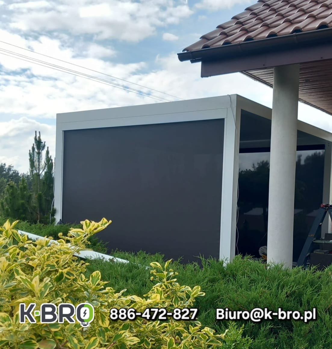 Nowoczesna pergola z szarą, zwijaną przesłoną, otoczona zielenią. Widoczne elementy konstrukcyjne i fragment dachu budynku. Ujęcie z poziomu gruntu.