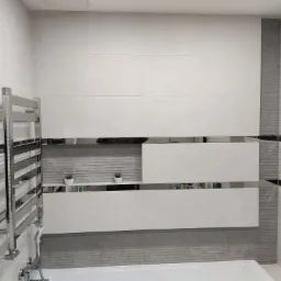 Wykończona łazienka z jasnymi, wielkoformatowymi płytkami, dekoracyjnymi wstawkami z lustrzanymi paskami, grzejnikiem drabinkowym i małymi roślinami doniczkowymi na półkach.