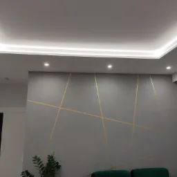 Szara ściana z geometrycznym wzorem w złotych liniach, podświetlany sufit z wbudowanymi punktowymi lampami, widoczne fragmenty zielonej rośliny doniczkowej i ciemnozielonego mebla.