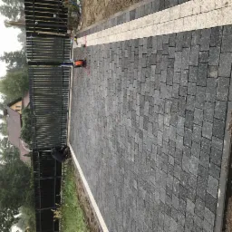 Ułożona szara kostka na podjeździe z białym pasem oddzielającym od czarnego metalowego ogrodzenia. Widoczny pracownik w trakcie pracy oraz zwinięty przewód elektryczny.