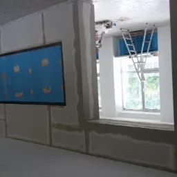 Wnętrze biura w trakcie remontu, ściany z płyt kartonowo-gipsowych przed wykończeniem, widoczna drabina przy oknie i fragment instalacji elektrycznej.