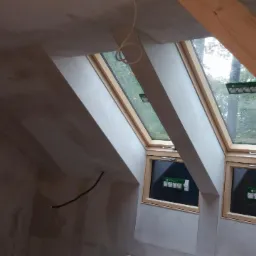 Poddasze w trakcie wykańczania, widoczne dwa okna dachowe z drewnianymi ramami, ściany pokryte świeżo nałożoną gładzią, drewniana konstrukcja dachu. Widoczne kable elektryczne wystające ze ściany.