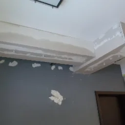 Fragment montażu podwieszanego sufitu z płyt kartonowo-gipsowych, widoczne profile i wstępne szpachlowanie łączeń na szarej ścianie.