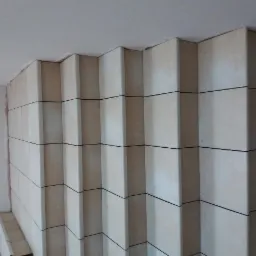 Ściana wyłożona beżowymi, wystającymi płytkami ceramicznymi z ciemnymi fugami, tworzącymi trójwymiarowy wzór schodkowy, widoczne niedociągnięcia na styku z sufitem i ścianą boczną.