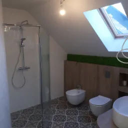 Nowoczesna łazienka na poddaszu z kabiną prysznicową, bidetem, toaletą i umywalką na drewnianym blacie, wykończona płytkami w stylu vintage i panelami drewnopodobnymi z mchem dekoracyjnym pod oknem...