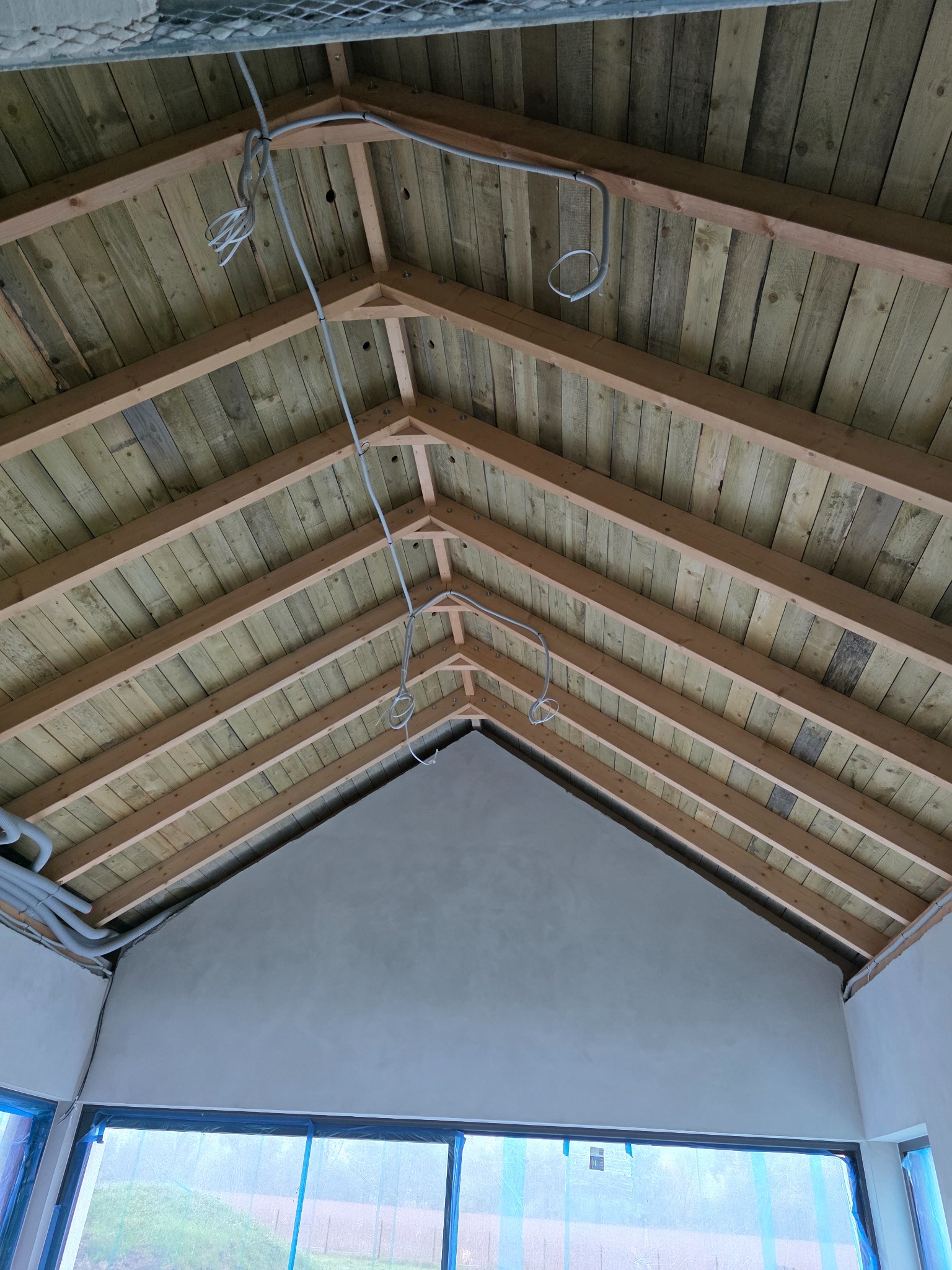 Drewniana podbitka dachowa z widocznymi kablami elektrycznymi, świeżo otynkowana ściana i okna zabezpieczone folią malarską. Wnętrze w trakcie wykańczania.