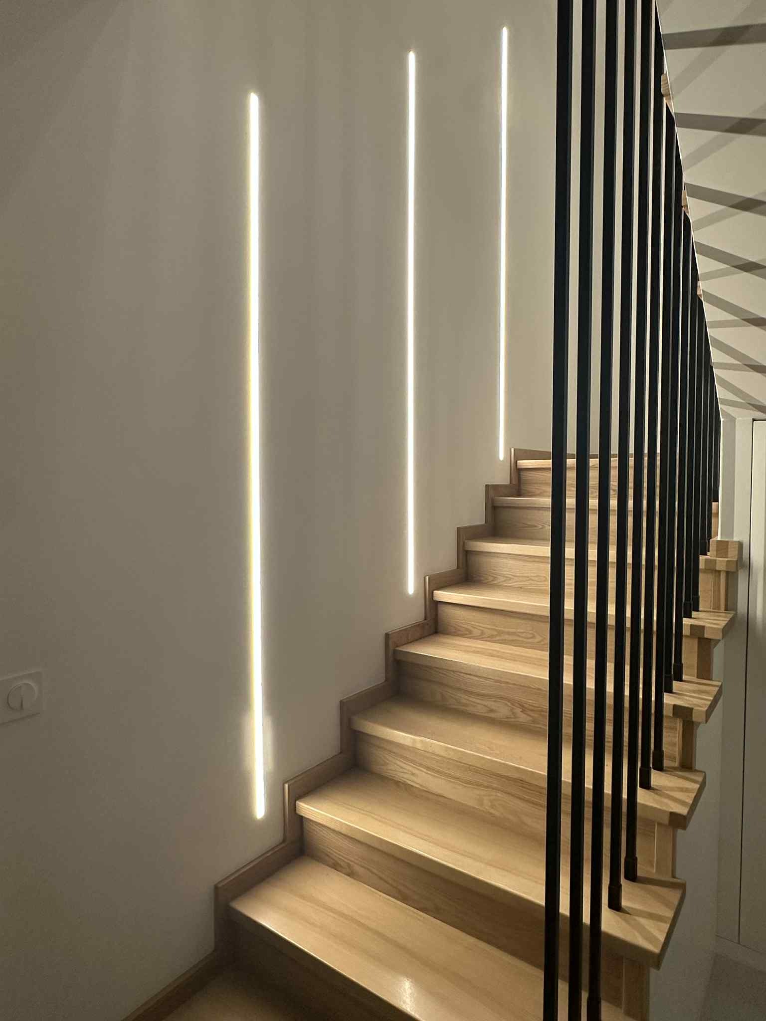 Nowoczesne schody z drewnianymi stopniami i czarną balustradą, oświetlone pionowymi pasami LED w ścianie, tworząc minimalistyczny i elegancki design.