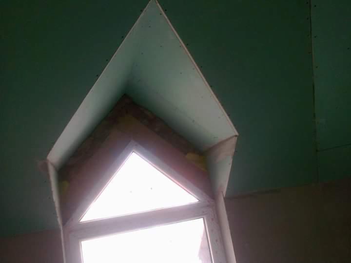 Montaż płyty gipsowo-kartonowej wokół okna o nietypowym, trójkątnym kształcie, widoczne łączenia i szpachlowanie, sufit i ściany częściowo pokryte zieloną płytą.