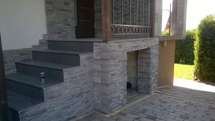 Zewnętrzne schody wejściowe wyłożone szarą płytką, z kamienną okładziną ściany i balustradą z metaloplastyki; na jednym ze stopni stoi puszka z napojem.