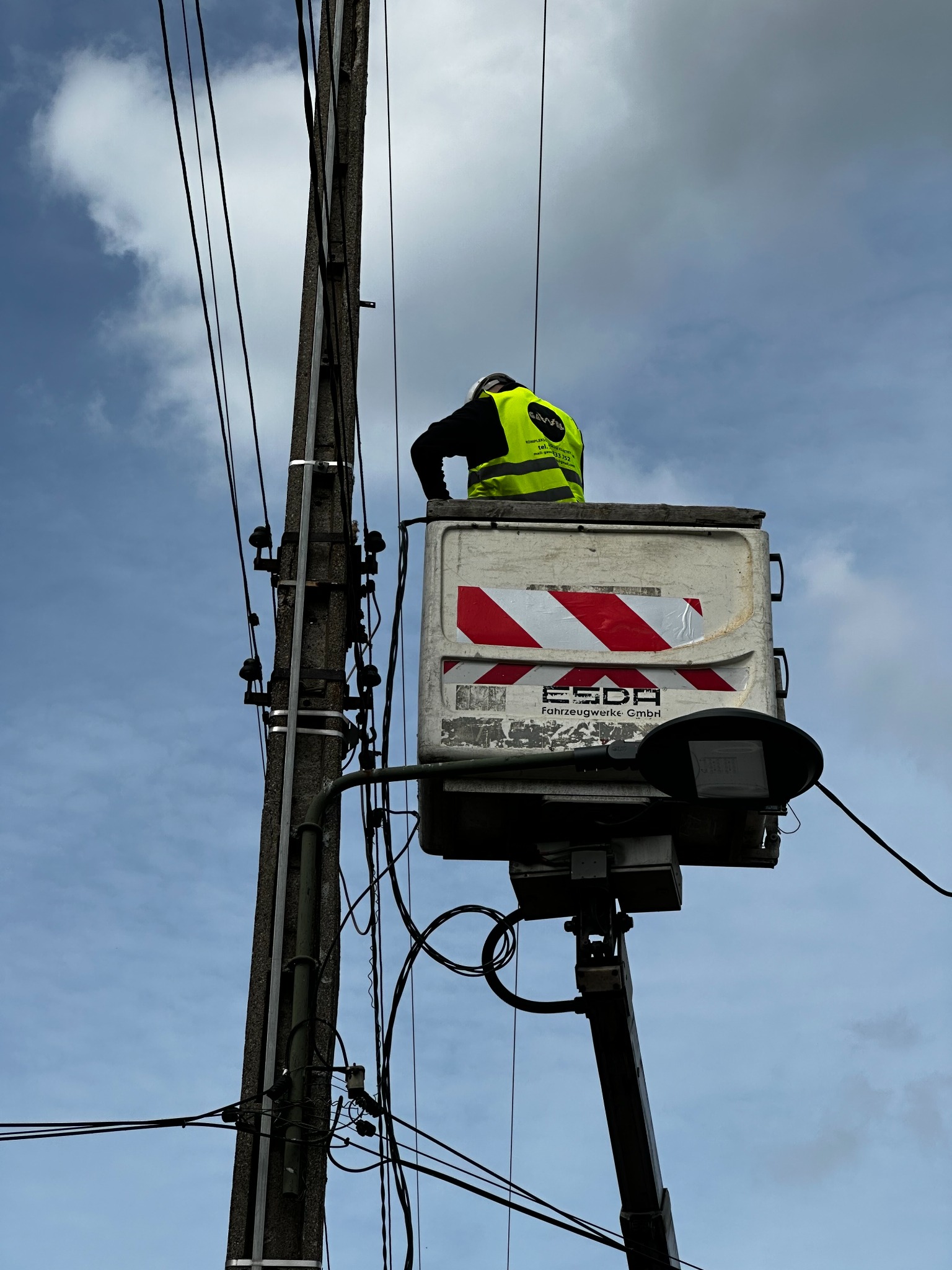 Pracownik w koszu podnośnika przy słupie energetycznym w Legionowie. Widoczne przewody i izolatory. Praca na wysokościach w żółtej kamizelce.