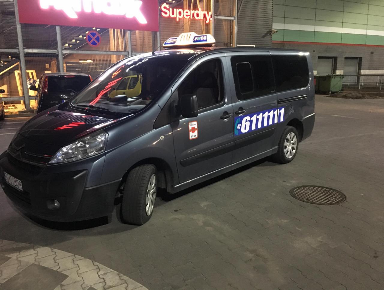 Szary van z numerem telefonu '6111111' na boku, zaparkowany przed budynkiem z szyldem 'TK Maxx' i 'Superdry'.
