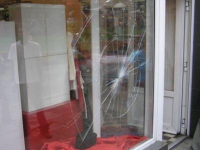 Rozbita szyba wystawowa sklepu odzieżowego w miejscowości ?. Pęknięcia układają się w pajęczynę z wyraźnym punktem uderzenia w centrum. Na pierwszym planie czerwony materiał, prawdopodobnie element...