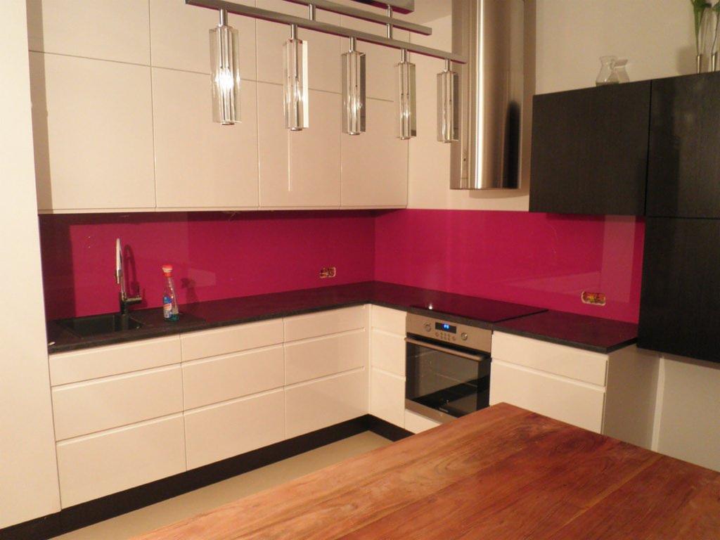 Nowoczesna kuchnia z białymi szafkami, czarnym blatem i intensywnie różowym panelem szklanym między blatem a szafkami wiszącymi, uzupełniona oświetleniem punktowym i drewnianym stołem na pierwszym...