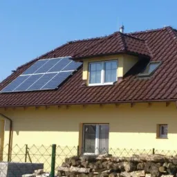 Żółty dom jednorodzinny z brązowym dachem, na którym zamontowano panele fotowoltaiczne; widoczne drewno opałowe przy ogrodzeniu.