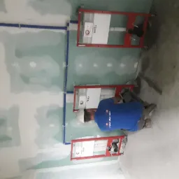 Instalacja podtynkowych stelaży WC marki TECE na tle surowych ścian z płyt kartonowo-gipsowych, widoczne niebieskie rury wodne i pracownik w niebieskiej koszulce podczas montażu.