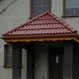 Zadaszenie nad wejściem do domu pokryte czerwoną dachówką ceramiczną, wsparte na ciemnych, ceglanych kolumnach. Widoczne okno na pierwszym piętrze i fragment rynny. Dekoracyjne wykończenie pod okapem.
