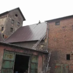 Pracownik na dachu krytym blachą trapezową, z drabiny opierającej się o ceglany budynek, w tle budynek z częściowo odpadającym tynkiem.