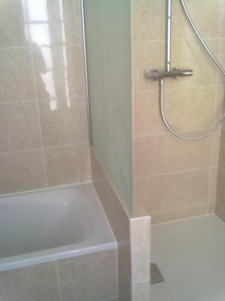 Beżowe płytki w łazience, widoczna wanna, kabina prysznicowa i bateria, z odpływem w rogu.