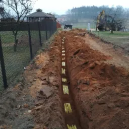 Wykop pod rury z widocznymi żółtymi osłonami, ziemia wokół, w tle ogrodzenie z siatki, altana, drzewa i koparka.