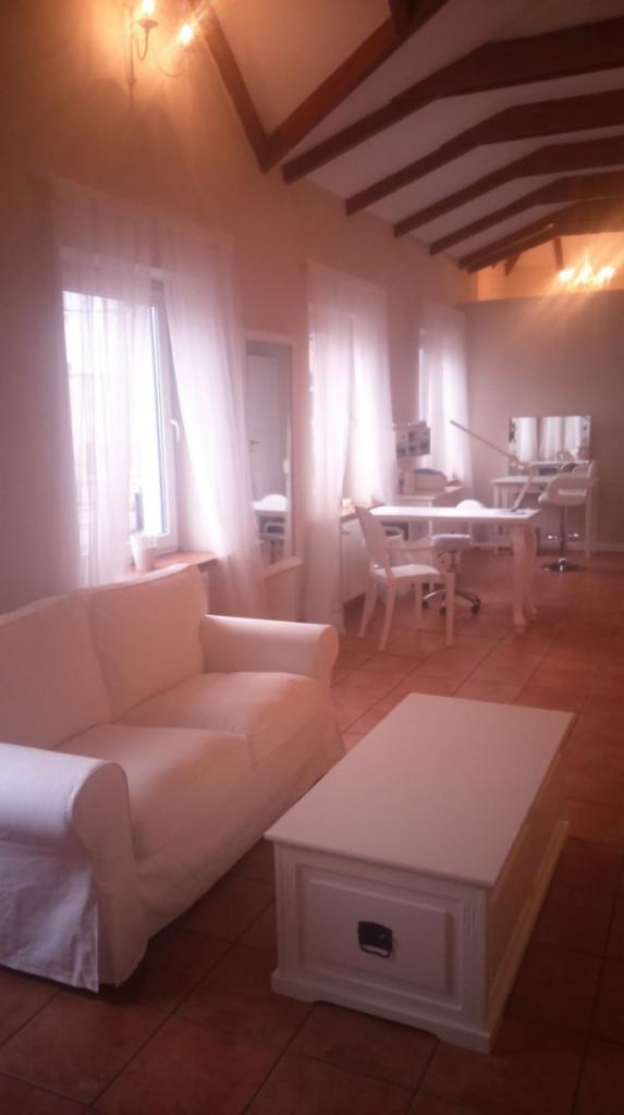 Jasne wnętrze salonu z białymi meblami, w tym sofą i stolikiem, oraz stanowiskami do zabiegów kosmetycznych w tle, oświetlone ciepłym światłem lamp.