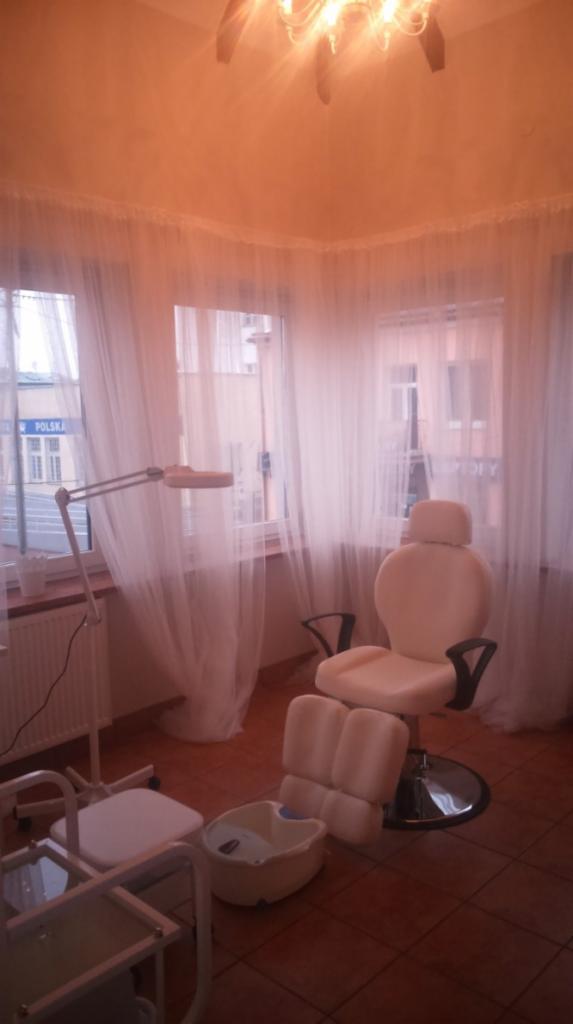 Jasne wnętrze gabinetu kosmetycznego z fotelem do pedicure, wanną do stóp i lampą powiększającą przy oknie z firankami.