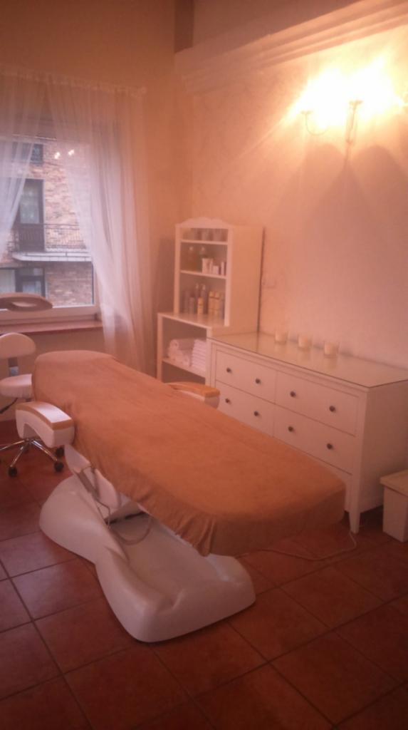 Jasne wnętrze salonu kosmetycznego z białym, regulowanym łóżkiem do zabiegów, komodą z szufladami i przeszklonym blatem oraz półkami z kosmetykami i ręcznikami, oświetlone ciepłym światłem