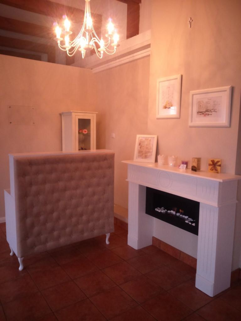 Jasne wnętrze salonu kosmetycznego z recepcją obitą materiałem, białym kominkiem dekoracyjnym i wiszącym żyrandolem.