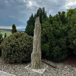 Ozdobna, wysoka kolumna z kamienia łupanego na betonowej podstawie, otoczona krzewami iglastymi i wysypana korą, z widokiem na zielone wzgórza pod pochmurnym niebem.