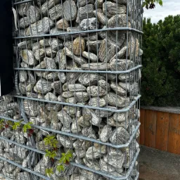 Gabion z szarego kamienia z widocznymi przewodami w peszlu, z zielenią pnącą się po siatce, na tle drewnianego ogrodzenia i iglaka.