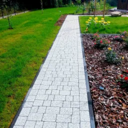 Ścieżka z jasnoszarej kostki brukowej z ciemną obwódką, biegnąca przez zadbany ogród z trawnikiem, kwiatami i korą drzewną.