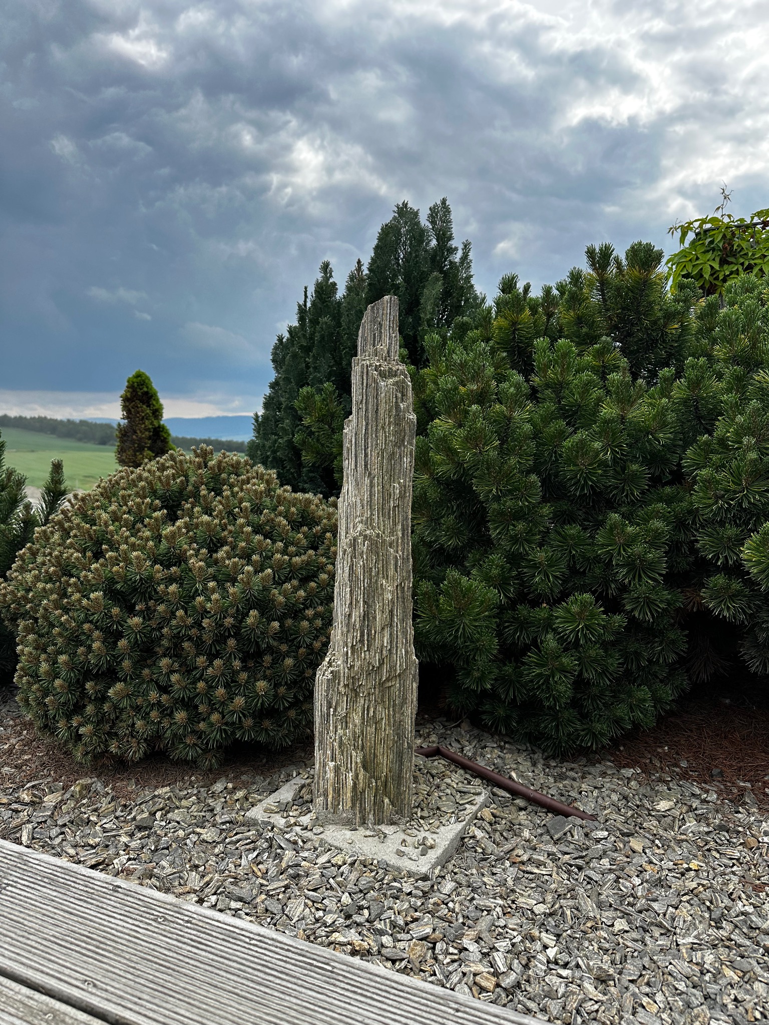 Ozdobna, wysoka kolumna z kamienia łupanego na betonowej podstawie, otoczona krzewami iglastymi i wysypana korą, z widokiem na zielone wzgórza pod pochmurnym niebem.