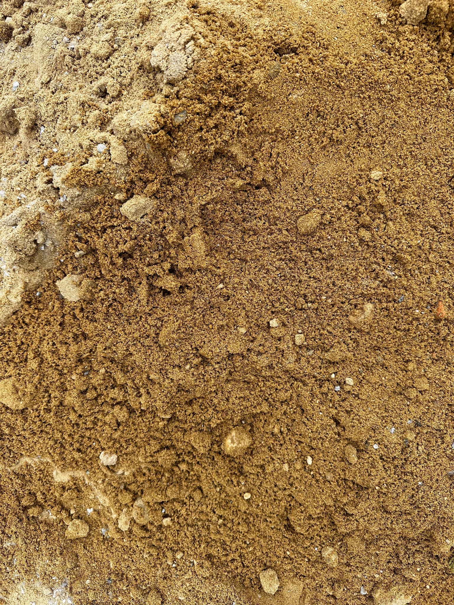 Góra piasku budowlanego o zróżnicowanej granulacji, od drobnego po grudki, z widocznymi kamykami i jaśniejszymi smugami na krawędziach.