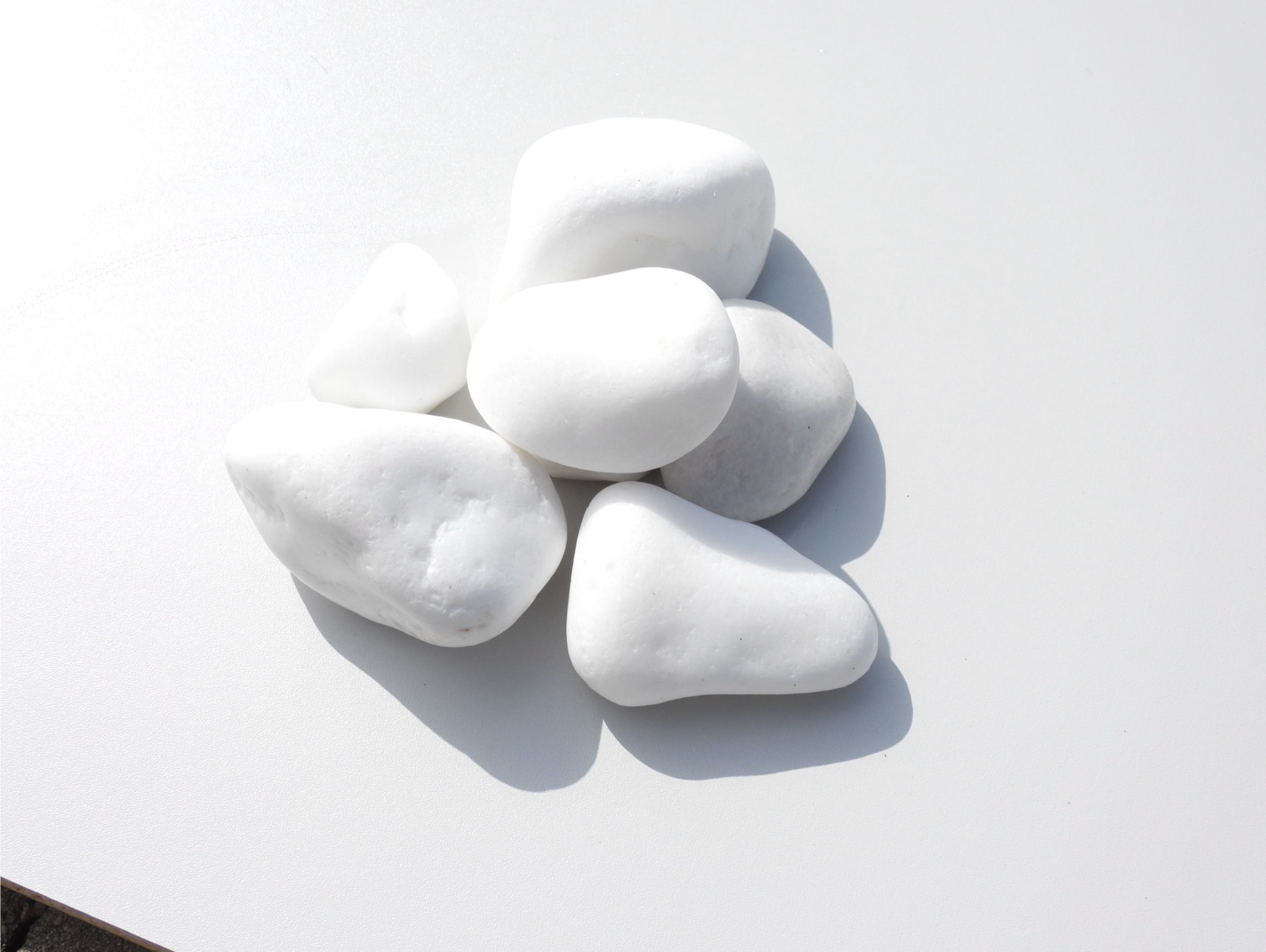 Białe, gładkie otoczaki ułożone na jasnej, matowej powierzchni w ostrym świetle słonecznym, rzucające wyraźne cienie.