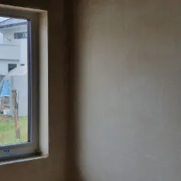 Świeżo nałożony tynk maszynowy na ścianie wewnętrznej, widok przez okno na budynek mieszkalny i konstrukcję foliową.