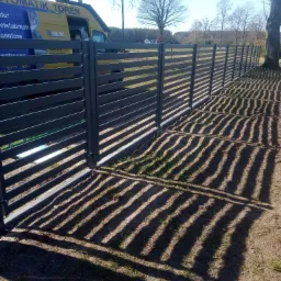 Długi, poziomy, szary płot panelowy z widocznym samochodem dostawczym w tle, rzucający wyraźny cień na ziemię, w tle drzewa bez liści i dom.