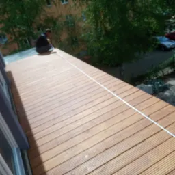 Montaż drewnianego tarasu na dachu budynku mieszkalnego, widoczny pracownik podczas układania desek, w tle zieleń drzew i zaparkowane samochody.