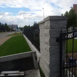 Fragment ogrodzenia z kutymi przęsłami i słupkami z szarej kostki, widok z wnętrza skrzyni ładunkowej samochodu dostawczego, automat do bramy w tle.