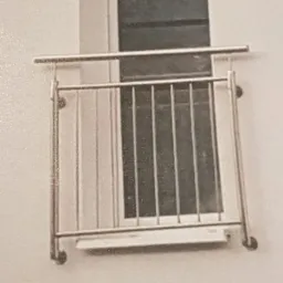 Niewielka balkonowa balustrada ze stali nierdzewnej, widoczna od zewnątrz, z prostymi pionowymi prętami, zamontowana przy oknie w jasnym budynku.