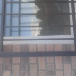 Czarna, stalowa krata okienna zamontowana na ceglanej ścianie budynku, widoczne śruby mocujące i odbicie nieba w szybie okiennej.