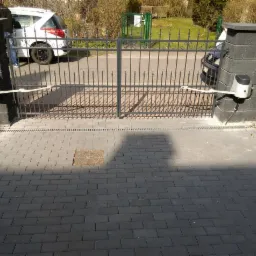 Dwuskrzydłowa brama wjazdowa z automatyką, zamontowana na murowanych słupkach z szarej kostki, widok z zewnątrz na posesję wyłożoną kostką brukową.