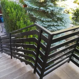 Ciemna, pozioma balustrada z metalowych profili na schodach zewnętrznych, widok z góry, w tle iglaki i pojemniki na śmieci.