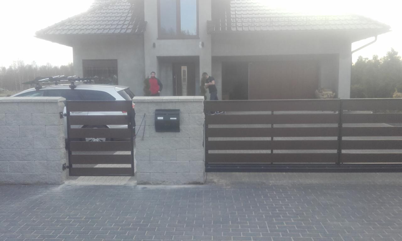 Nowoczesna posesja z przesuwną bramą i furtką z poziomych paneli, murowane słupki z szarej kostki, skrzynka na listy, samochód z bagażnikiem dachowym widoczny za furtką, dom w tle, kostka brukowa...