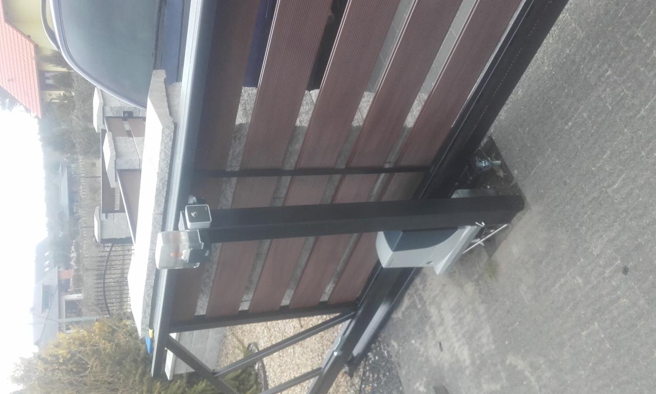 Szczegółowy widok boczny bramy przesuwnej z brązowymi panelami kompozytowymi i czarną ramą, widoczny mechanizm automatyki z szarą obudową, betonowa podstawa i częściowo widoczny samochód w tle.