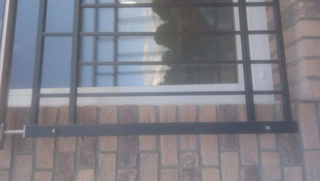 Czarna, stalowa krata okienna zamontowana na ceglanej ścianie budynku, widoczne śruby mocujące i odbicie nieba w szybie okiennej.