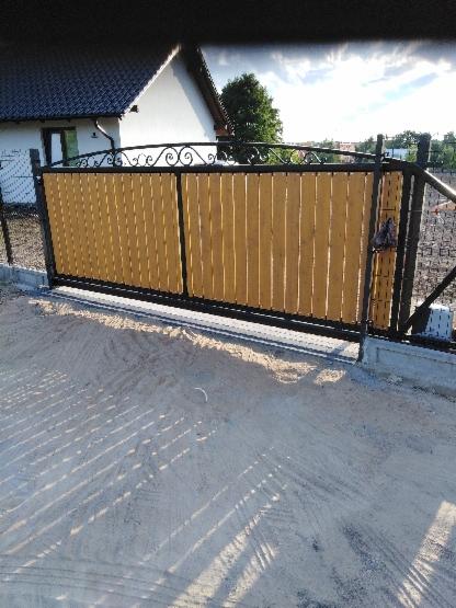 Nowoczesna brama wjazdowa z drewnianym wypełnieniem i czarnymi, kutymi elementami dekoracyjnymi na górze, osadzona na betonowej podmurówce, z widocznym fragmentem domu w tle.