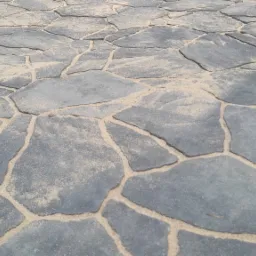 Nierówna powierzchnia z szarych, nieregularnych płyt kamiennych, wypełniona piaskiem, tworząca ścieżkę lub podjazd.
