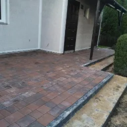 Ułożona kostka brukowa w odcieniach brązu i szarości, tworząca taras przed budynkiem z ciemnymi drzwiami i jasną elewacją, z dwoma stopniami wykończonymi szarą kostką i widoczną ziemią po bokach...