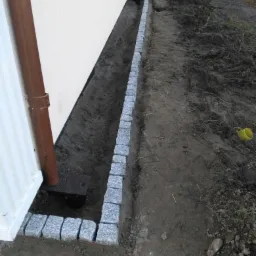 Granitowa obwódka wzdłuż ściany domu, z brązową rurą spustową, otoczona świeżo przekopaną ziemią.