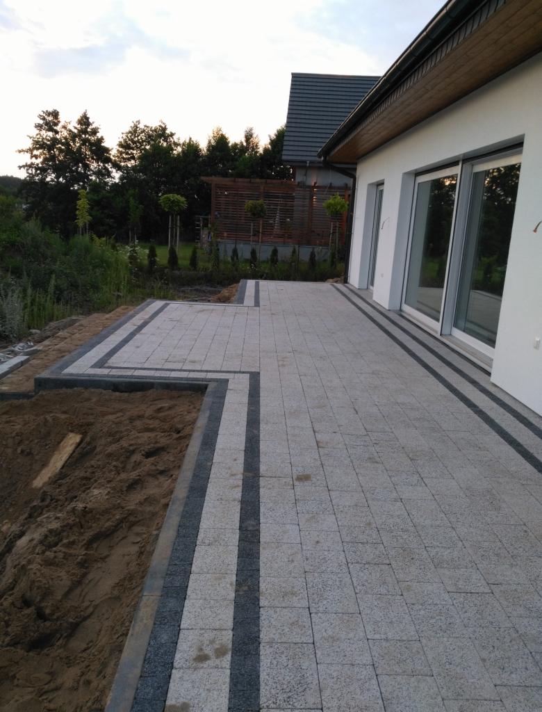 Ułożona szara kostka brukowa z ciemnymi pasami obramowania, przylegająca do białego budynku z dużymi oknami przesuwnymi, obok wykopana ziemia przygotowana pod dalsze prace.